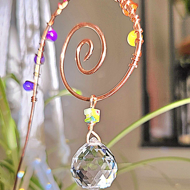 Close-up of a handmade plant stake suncatcher for garden decor, crafted with 16-gauge copper wire in a spiral design. The stake is adorned with colorful glass beads and features a millefiori glass bead hanging above a 20mm K9 crystal ball. The crystal sparkles in the sunlight, scattering rainbow light effects.