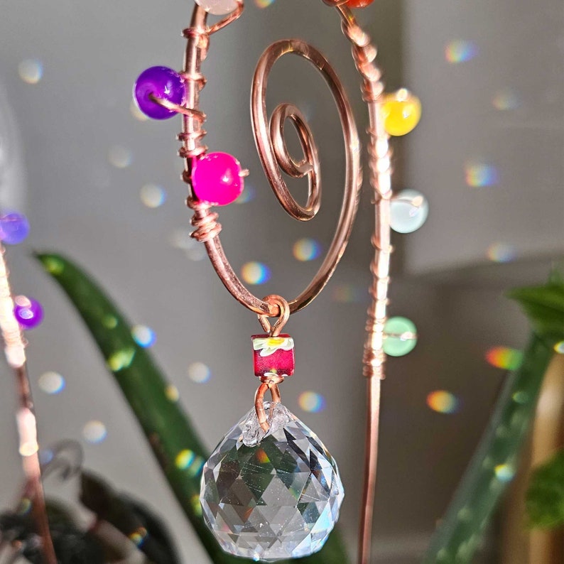 Close-up of a handmade plant stake suncatcher for garden decor, crafted with 16-gauge copper wire in a spiral design. The stake is adorned with colorful glass beads and features a millefiori glass bead hanging above a 20mm K9 crystal ball. The crystal sparkles in the sunlight, scattering rainbow light effects.