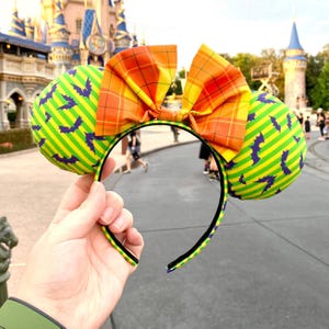 May include: A headband featuring two round ears with a lime green and yellow striped pattern decorated with small purple bats. A large, central bow sits between the ears, displaying an orange and yellow plaid pattern. The headband frame is black.