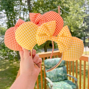 May include: A headband featuring two large, padded circular ears covered in an orange and yellow gingham check fabric. A large, matching fabric bow is centered between the ears, also featuring the same orange and yellow checkered pattern.