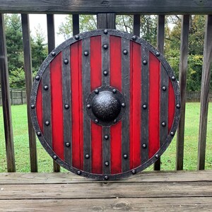 Puede incluir: Un escudo redondo de madera con un dise&ntilde;o rojo y gris. El escudo tiene rayas rojas verticales separadas por bandas met&aacute;licas grises. Un jefe central de metal texturizado y un borde met&aacute;lico completan el dise&ntilde;o. El escudo tiene aproximadamente 76 cm de di&aacute;metro.