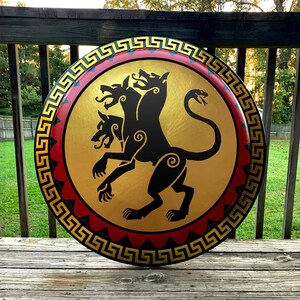 May include: A round, wooden shield with a black and gold design. The shield features a three-headed dog with a snake tail, a Greek key border, and red and gold accents.