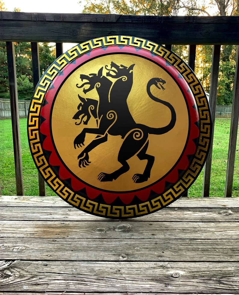 May include: A round, wooden shield with a black and gold design. The shield features a three-headed dog with a snake tail, a Greek key border, and red and gold accents.