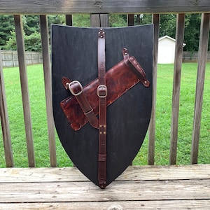May include: A black wooden shield with a brown leather strap and a brown leather quiver attached. The shield is a teardrop shape. The leather has silver buckles and rivets. The shield is displayed on a wooden deck.