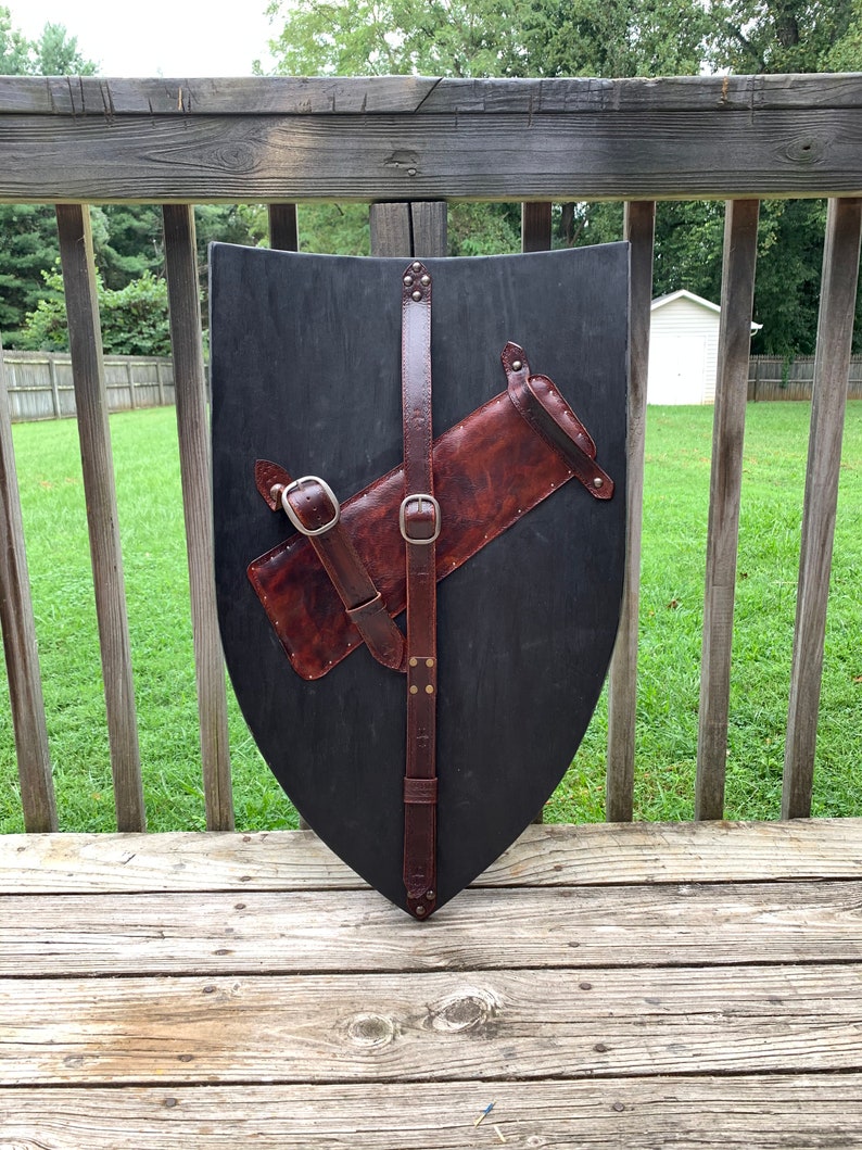 May include: A black wooden shield with a brown leather strap and a brown leather quiver attached. The shield is a teardrop shape. The leather has silver buckles and rivets. The shield is displayed on a wooden deck.