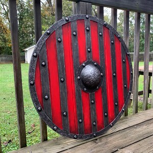 Puede incluir: Un escudo vikingo redondo con un dise&ntilde;o a rayas rojas y negras. El escudo tiene un borde met&aacute;lico y un centro abultado. Fabricado en madera y metal, est&aacute; dise&ntilde;ado para la protecci&oacute;n en combate.