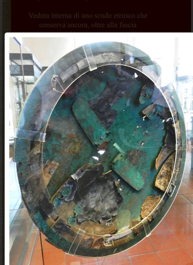 May include: A close-up view of the inside of an Etruscan shield. The shield is made of bronze and is decorated with a geometric pattern. The shield is in a museum display case.
