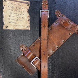 May include: A black wooden shield with brown leather straps and accents. The shield has a scroll with the text "Take up the shield of faith, with which you can extinguish all the flaming arrows of the evil one. Ephesians 6:16."