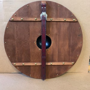 Puede incluir: Un escudo redondo de madera con acabado marr&oacute;n oscuro, remaches de lat&oacute;n y un jefe central de metal negro. Una correa de cuero burdeos con detalles met&aacute;licos plateados y dise&ntilde;os grabados est&aacute; unida al escudo.