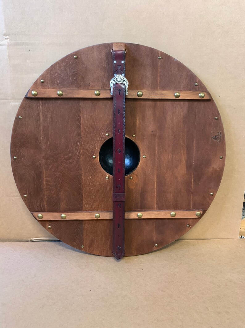 Puede incluir: Un escudo redondo de madera con acabado marr&oacute;n oscuro, remaches de lat&oacute;n y un jefe central de metal negro. Una correa de cuero burdeos con detalles met&aacute;licos plateados y dise&ntilde;os grabados est&aacute; unida al escudo.