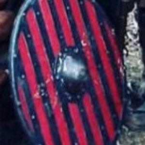 Puede incluir: Un escudo redondo con un borde negro y un patr&oacute;n de rayas verticales rojas. El escudo tiene un jefe central de metal y probablemente se utiliza para la protecci&oacute;n en combate o recreaciones hist&oacute;ricas.
