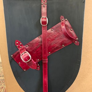May include: A black shield with a curved top and a pointed bottom. It features red leather straps and accents, including a central vertical strap with a buckle and a diagonal strap. The shield is adorned with silver rivets.