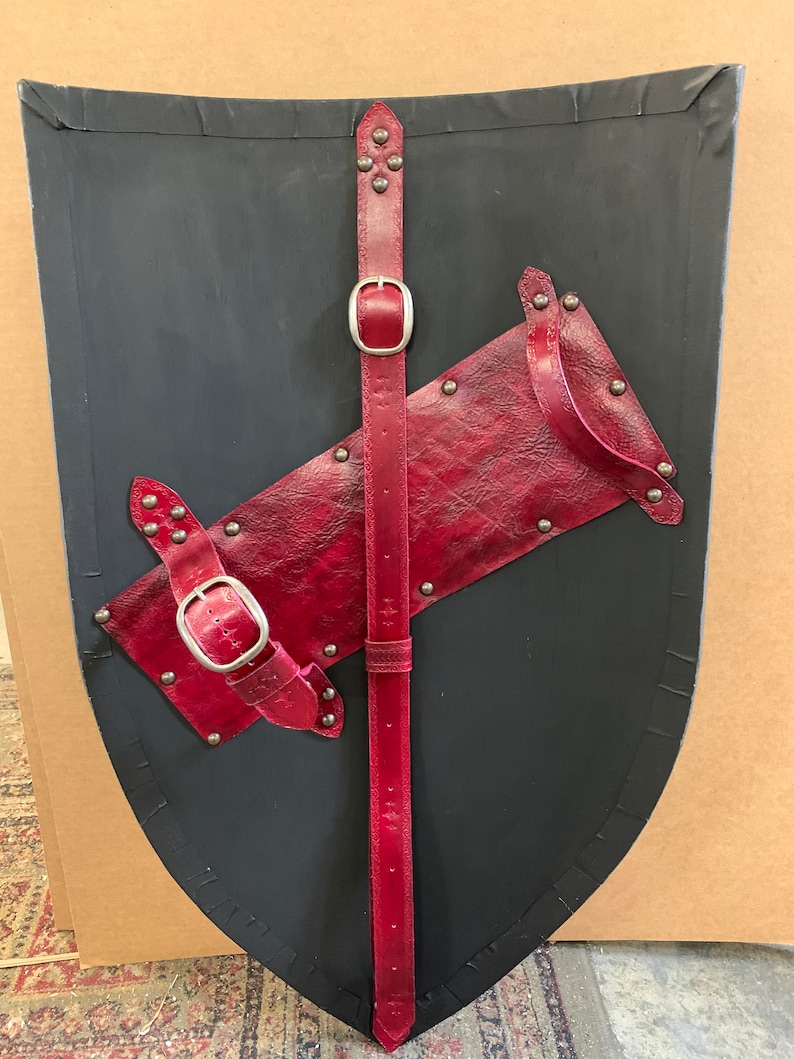 May include: A black shield with a curved top and a pointed bottom. It features red leather straps and accents, including a central vertical strap with a buckle and a diagonal strap. The shield is adorned with silver rivets.