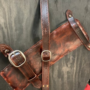 May include: A brown leather quiver and belt set with decorative embossed patterns and silver buckles. The quiver has a rectangular shape and is attached to a long belt with a buckle. The belt has a series of rivets and a cross design.