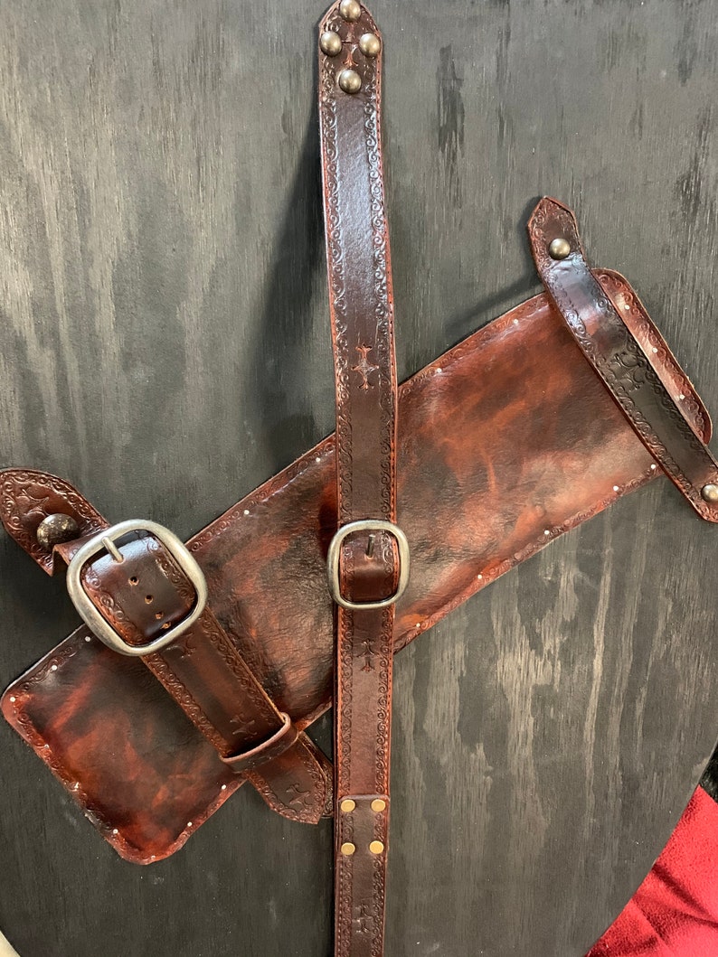 May include: A brown leather quiver and belt set with decorative embossed patterns and silver buckles. The quiver has a rectangular shape and is attached to a long belt with a buckle. The belt has a series of rivets and a cross design.