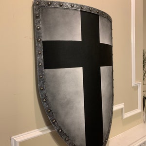 May include: A medieval-style shield with a silver and black design. The shield features a large black cross on a silver background, with a riveted metal border. The shield is mounted on a wall.
