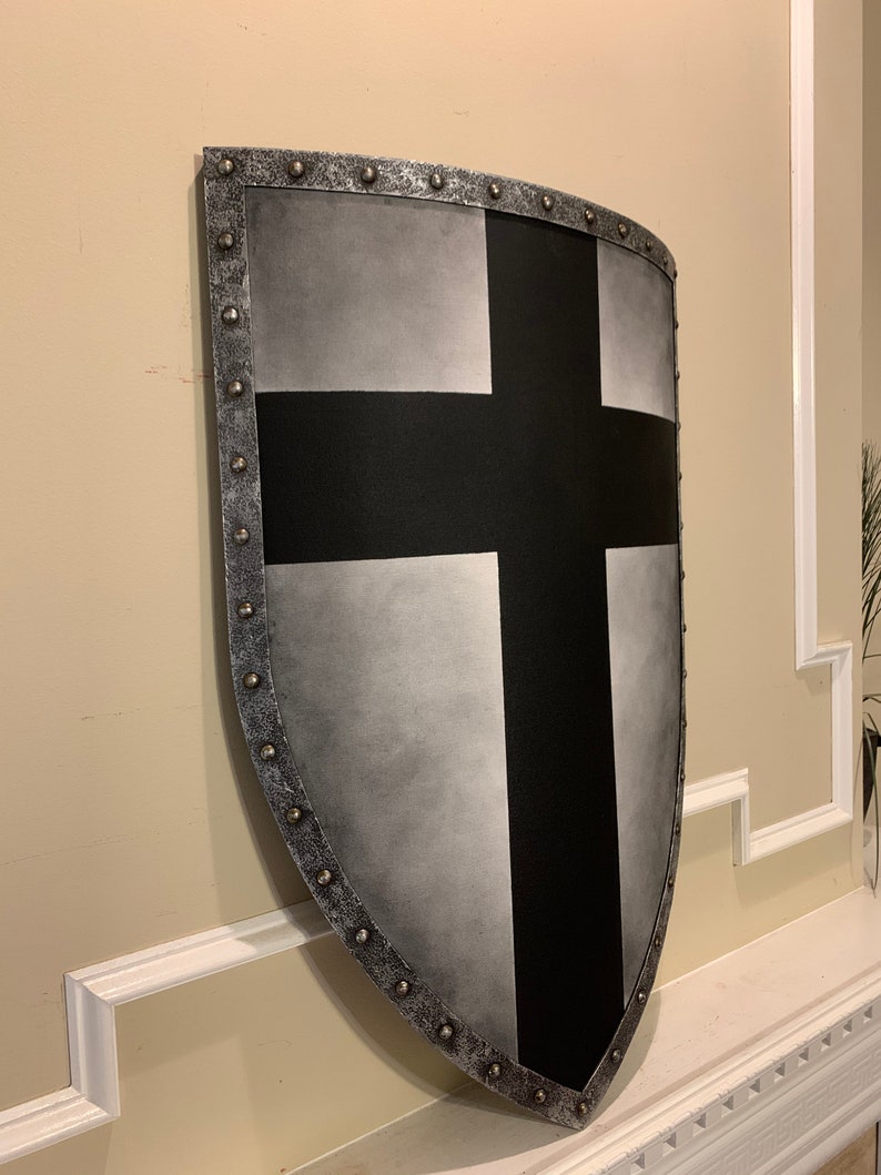 May include: A medieval-style shield with a silver and black design. The shield features a large black cross on a silver background, with a riveted metal border. The shield is mounted on a wall.