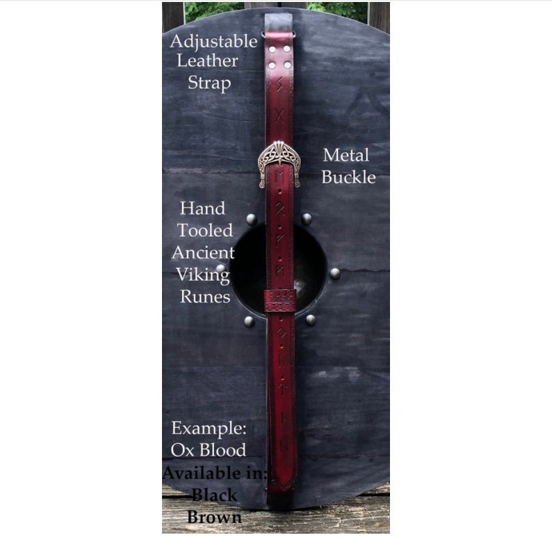 Puede incluir: Un escudo redondo de madera oscura con un agujero central y una correa de cuero burdeos. La correa presenta runas vikingas hechas a mano y una hebilla de metal. El texto indica que la correa es ajustable y est&aacute; disponible en negro o marr&oacute;n.