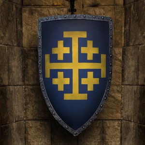 May include: A medieval-style shield with a dark blue background and a gold Jerusalem cross design. The shield has a silver trim with rivets and is mounted on a stone wall. A candle holder with lit candles is above the shield.
