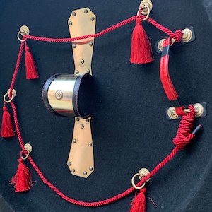 May include: A black round shield with a brass plate and red tassels attached to a red rope. The shield has a leather strap on the back.