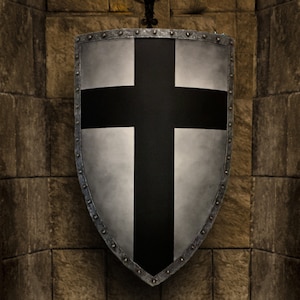 May include: A silver shield with a black cross design, mounted on a stone wall. The shield has a riveted border and a slightly weathered appearance. A wrought iron candle holder with lit candles is above the shield, casting a warm glow.