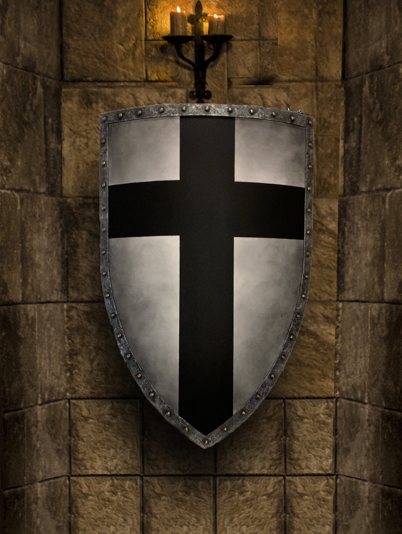 May include: A silver shield with a black cross design, mounted on a stone wall. The shield has a riveted border and a slightly weathered appearance. A wrought iron candle holder with lit candles is above the shield, casting a warm glow.