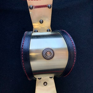 May include: A gold metal cuff bracelet with a black leather band and red stitching. The band has a Greek key design and a circular metal accent with a sunburst design.