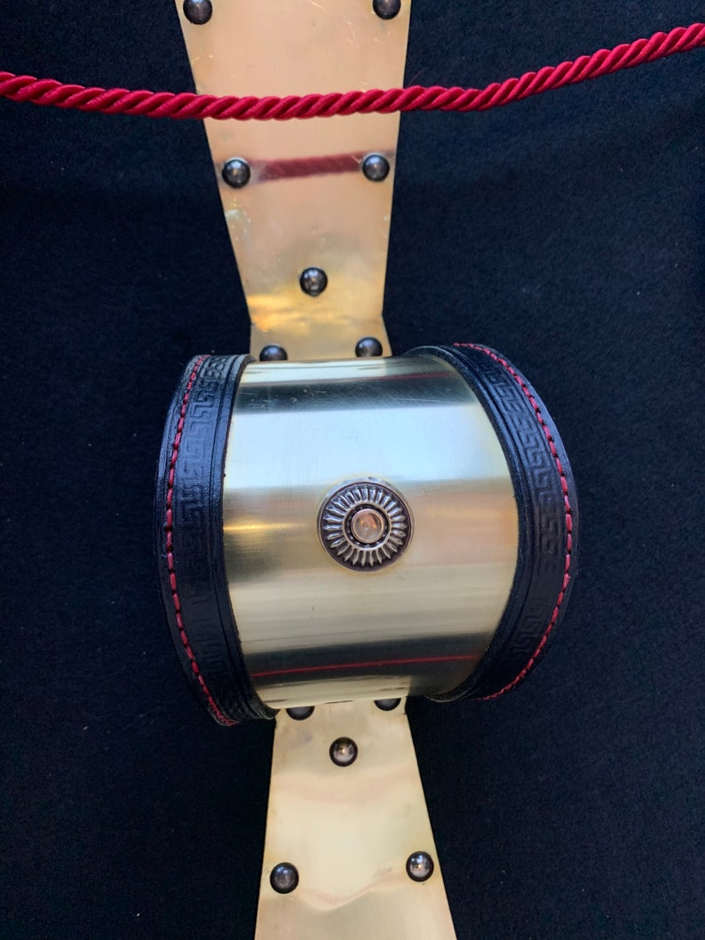 May include: A gold metal cuff bracelet with a black leather band and red stitching. The band has a Greek key design and a circular metal accent with a sunburst design.