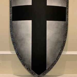 May include: A shield-shaped wall hanging with a black cross design. The shield has a silver-colored border with rivets and a gradient gray background.
