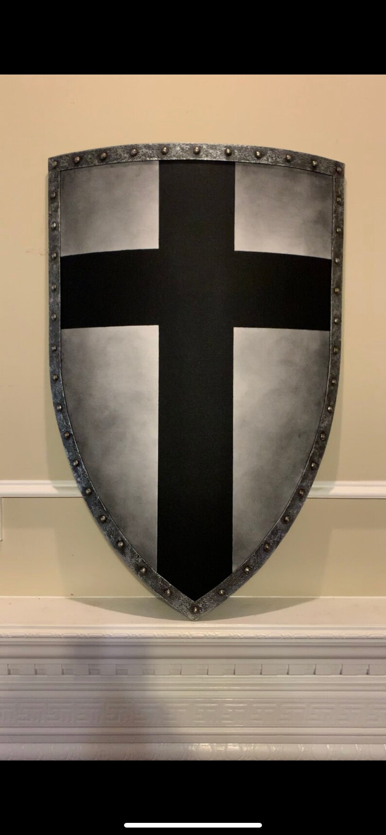 May include: A shield-shaped wall hanging with a black cross design. The shield has a silver-colored border with rivets and a gradient gray background.