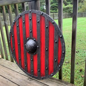 Puede incluir: Un escudo redondo de madera con un dise&ntilde;o a rayas rojas y negras. El escudo tiene un borde de metal y un centro abultado. El escudo mide aproximadamente 76 cm de di&aacute;metro, con tachuelas de metal y un aspecto envejecido.