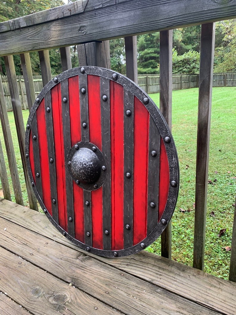 Puede incluir: Un escudo redondo de madera con un dise&ntilde;o a rayas rojas y negras. El escudo tiene un borde de metal y un centro abultado. El escudo mide aproximadamente 76 cm de di&aacute;metro, con tachuelas de metal y un aspecto envejecido.