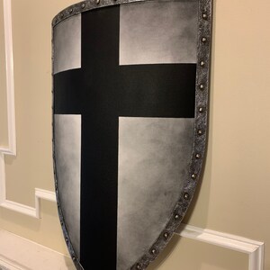 May include: A medieval-style shield with a black cross design on a silver background. The shield has a curved shape and is trimmed with metal studs around the edges. The shield is mounted on a wall.