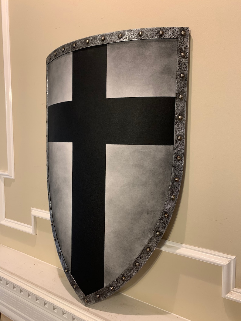 May include: A medieval-style shield with a black cross design on a silver background. The shield has a curved shape and is trimmed with metal studs around the edges. The shield is mounted on a wall.