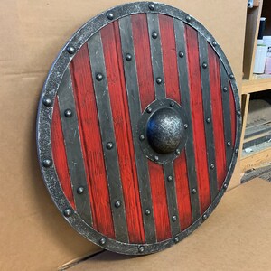 Puede incluir: Escudo de madera redondo con un dise&ntilde;o rojo y gris. El escudo presenta tablas de madera rojas verticales, bandas de metal grises y un jefe central de metal. El borde exterior est&aacute; adornado con una banda de metal gris y remaches.