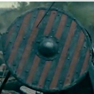 Puede incluir: Escudo redondo de madera con un patr&oacute;n de rayas rojas y azul oscuro. El escudo tiene tachuelas met&aacute;licas alrededor del borde y un saliente central. Dise&ntilde;o vikingo.