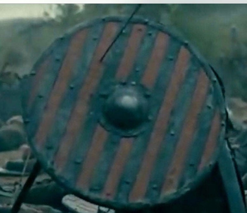 Puede incluir: Escudo redondo de madera con un patr&oacute;n de rayas rojas y azul oscuro. El escudo tiene tachuelas met&aacute;licas alrededor del borde y un saliente central. Dise&ntilde;o vikingo.