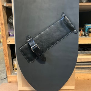 May include: A black shield with a curved top and pointed bottom. A black leather strap with a buckle and rivets is attached diagonally across the shield. The shield appears to be made of wood or a similar material.