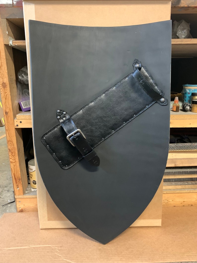 May include: A black shield with a curved top and pointed bottom. A black leather strap with a buckle and rivets is attached diagonally across the shield. The shield appears to be made of wood or a similar material.