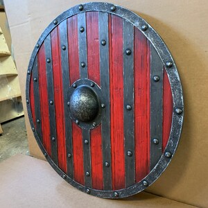 Puede incluir: Un escudo redondo de madera con un esquema de color rojo y negro. El escudo tiene tablas de madera rojas verticales, acentuadas por bandas y remaches de metal oscuro. Un jefe central de metal y un borde de metal completan el dise&ntilde;o.