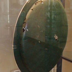 May include: A large, round, green metal plate with a worn surface. The plate has several small chips and scratches on its surface. The plate is displayed in a glass case.
