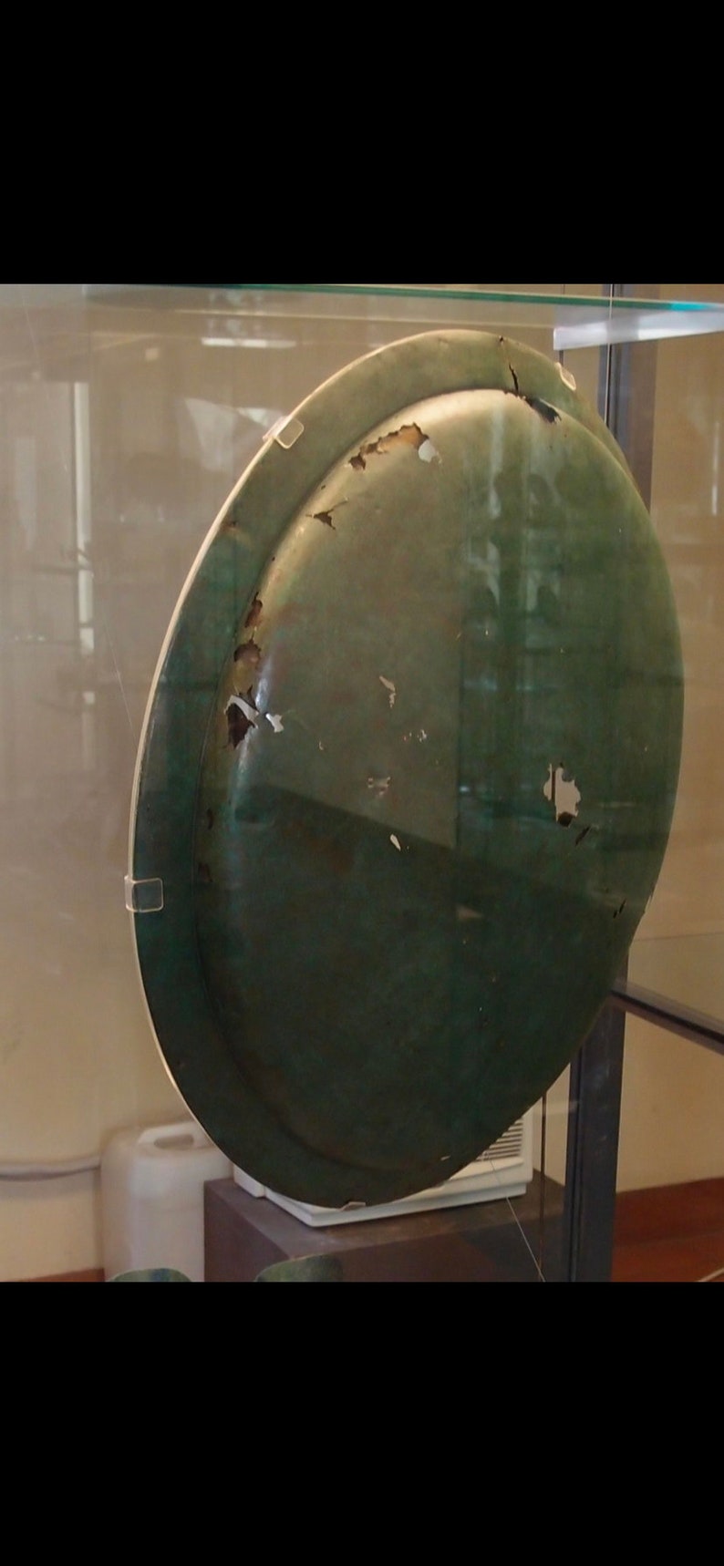 May include: A large, round, green metal plate with a worn surface. The plate has several small chips and scratches on its surface. The plate is displayed in a glass case.