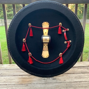 May include: A round, black shield with a gold metal center and red tassels. The shield is decorated with a red rope and gold metal accents.