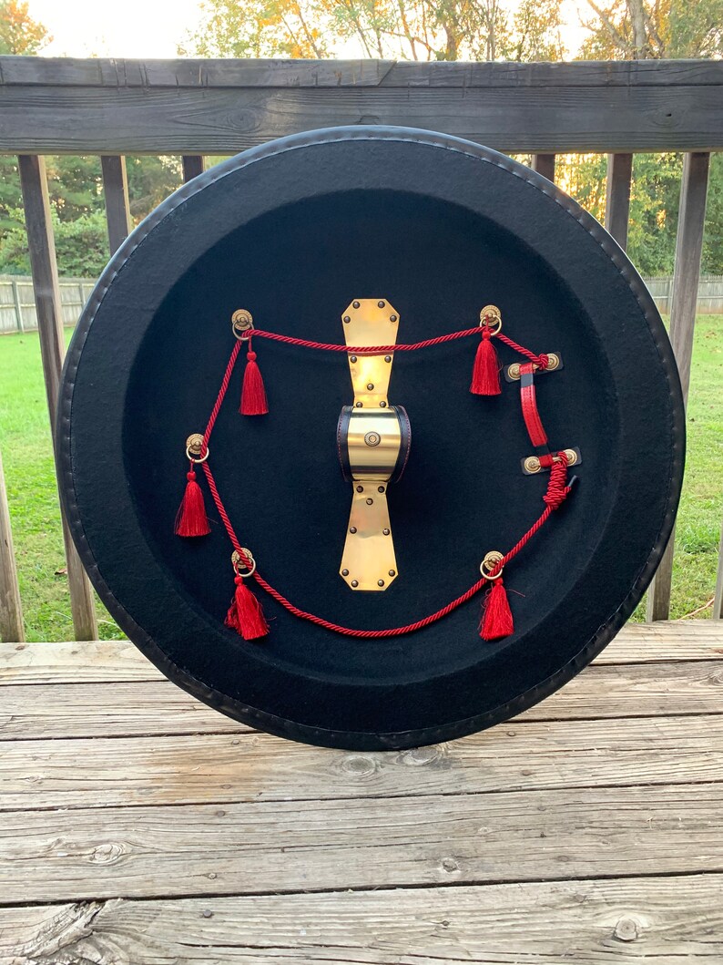 May include: A round, black shield with a gold metal center and red tassels. The shield is decorated with a red rope and gold metal accents.