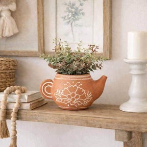 May include: A terracotta teapot-shaped planter featuring a white hand-painted floral design on the front. The planter contains a small green leafy plant and is positioned on a wooden surface next to a stack of books and a white pillar candle.