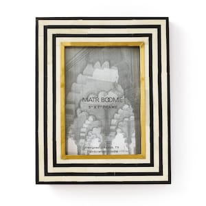 May include: A rectangular photo frame with a black and white striped border and a gold inner frame. The frame holds a black and white image. The frame is 12.7cm x 17.8cm. Text on the frame reads "MATR BOOMIE 5" X 7" FRAME Designed in Austin, TX Handcrafted in India."