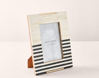 May include: A rectangular picture frame with a 4 inch by 6 inch opening. The frame features a two-tone design with a cream-colored upper section and a lower section composed of alternating horizontal black and cream stripes. Text reads MATR BOOMIE 4 X 6 FRAME.