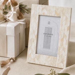 Puede incluir: Un marco de fotos rectangular con incrustaciones de patrón de espiga en color crema y blanco roto. El marco contiene una tarjeta con el texto MATR BOOMIE 5" X 7" FRAME y una ilustración de una puerta. El marco tiene orientación vertical.