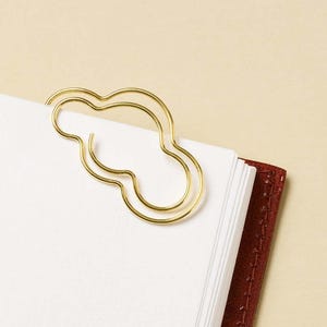 Puede incluir: Un clip de metal dorado con forma de contorno de nube estilizada. El clip está sujeto en la esquina superior de un montón de hojas de papel blanco, que descansan contra una cubierta de cuaderno de cuero de color marrón rojizo oscuro.