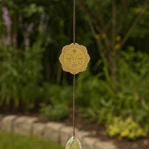 May include: A hanging suncatcher featuring a circular brass-toned metal charm with an engraved sun face design, suspended by a thin metal chain from a circular ring, with a clear, faceted, teardrop-shaped glass crystal hanging at the bottom.